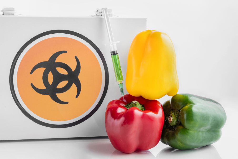 A black and orange symbol signifying that toxic chemicals are near is posted behind a syringe full of green liquid. The syringe is injecting into a red bell pepper at next to a yellow and green bell pepper, perhaps representing a call to prevention of cancer causing chemicals in food.