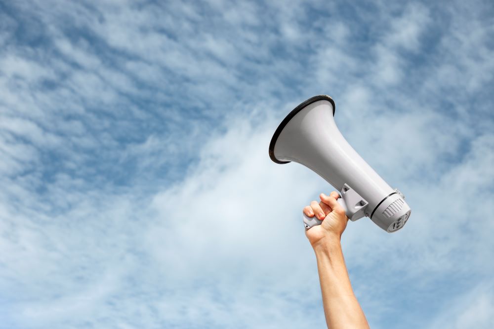 Single arm raises megaphone up against a partly cloudy sky, perhaps to help organize volunteers at a nonprofit organization's event or spread the word about the nonprofit's mission.