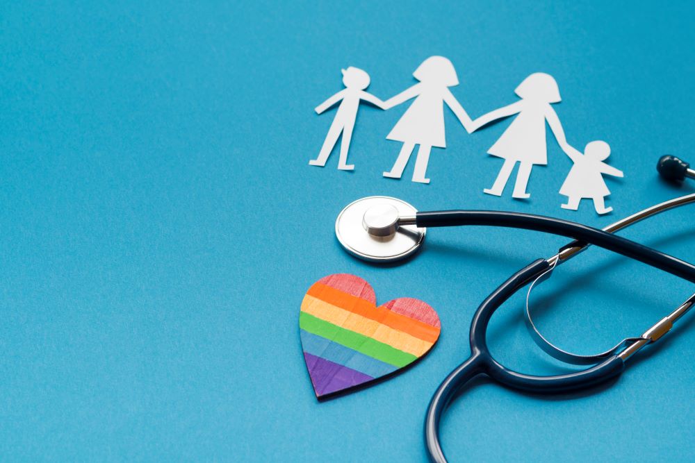 Image of a stethoscope resting over a rainbow heart and a paper cut-out family with two moms represents the importance of gay friendly health care and LGBTQ+ affirming doctors in gay family planning and gay healthcare support.