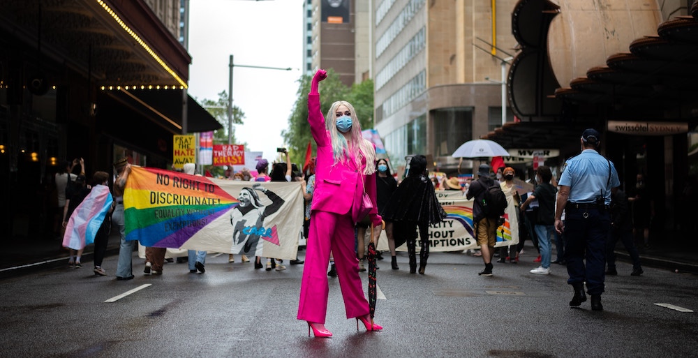 An individual in a hot pink suit stands in front of a crowd at a pride parade, fist raised, or perhaps it's a bi visibility month celebration. 