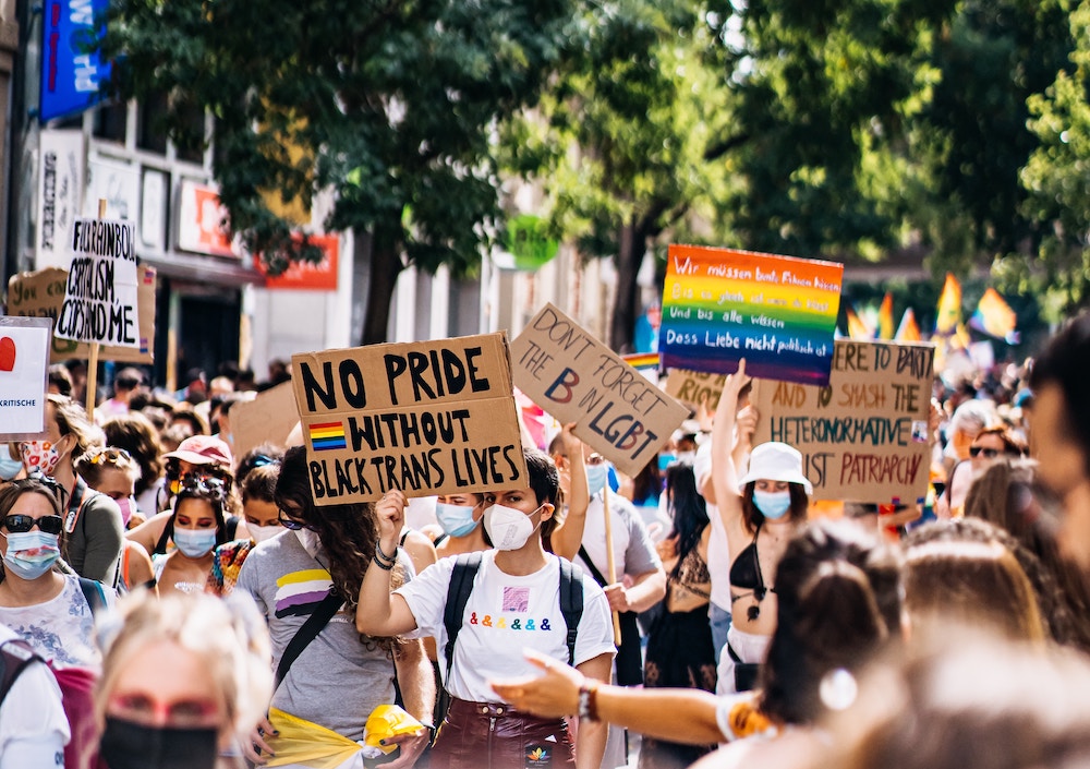 Many people gather holding banners and signs in support of LGBTQ+ individuals, bi awareness and visibility, and the rest of the queer community at a pride parade or a bi visibility month march.