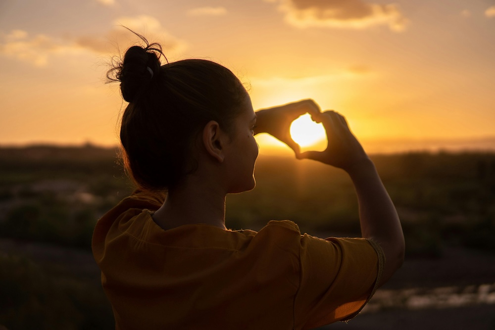 Joyful woman at sunset smiling and forming a heart with her hands, celebrating positivity, love, and the beauty of the golden hour.