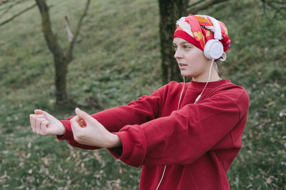 A woman is standing in a sunny park, stretching one arm overhead while holding her other arm for support. She is wearing a colorful head wrap, wireless earbuds, and athletic wear, including a tank top and leggings. She appears relaxed and focused, enjoying her music as she stretches against a backdrop of green grass and trees.