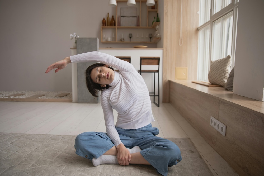 A woman sits cross-legged on a yoga mat, wearing cozy, loose-fitting clothes. She stretches one arm overhead and leans gently to the side, elongating her torso. Her other hand rests on the mat for support. The serene indoor setting features soft lighting and a calm atmosphere, complementing her relaxed posture.