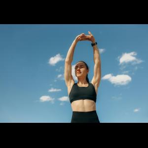 A woman in workout clothes is outdoors on a bright, sunny day with a vibrant blue sky overhead. She is mid-stretch, with one arm reaching high above her head and the other resting on her hip. She is wearing a tank top and leggings, with her hair tied back, and she appears energized and focused.