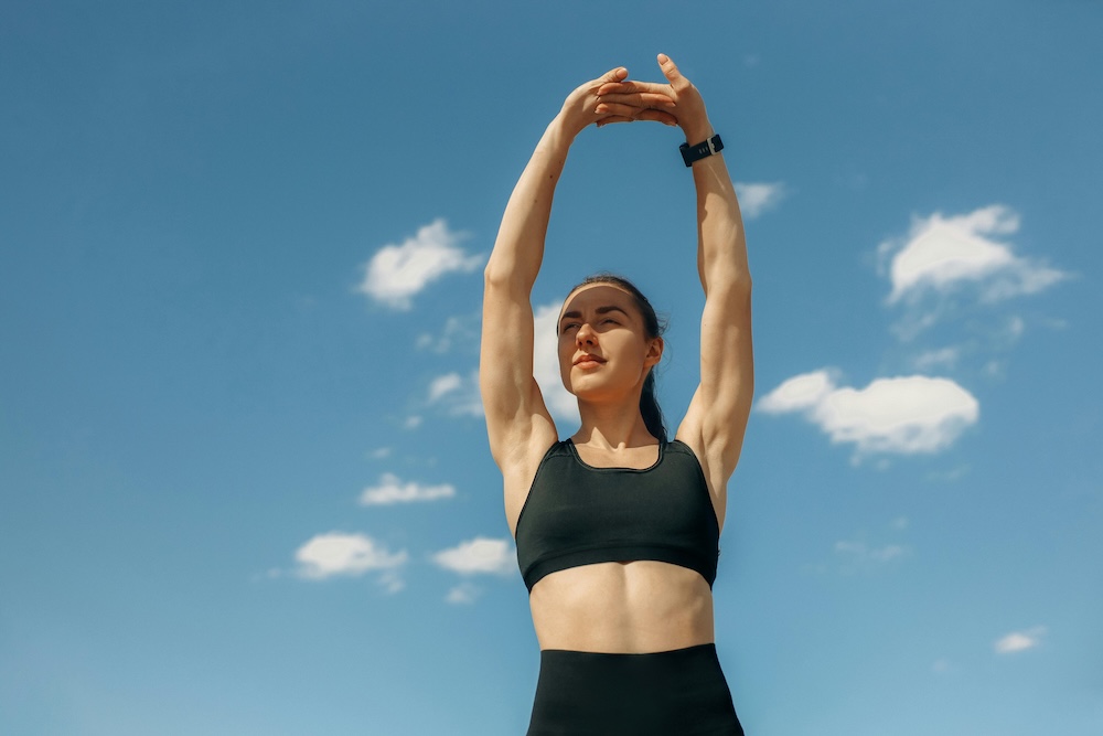 A woman in workout clothes is outdoors on a bright, sunny day with a vibrant blue sky overhead. She is mid-stretch, with one arm reaching high above her head and the other resting on her hip. She is wearing a tank top and leggings, with her hair tied back, and she appears energized and focused.