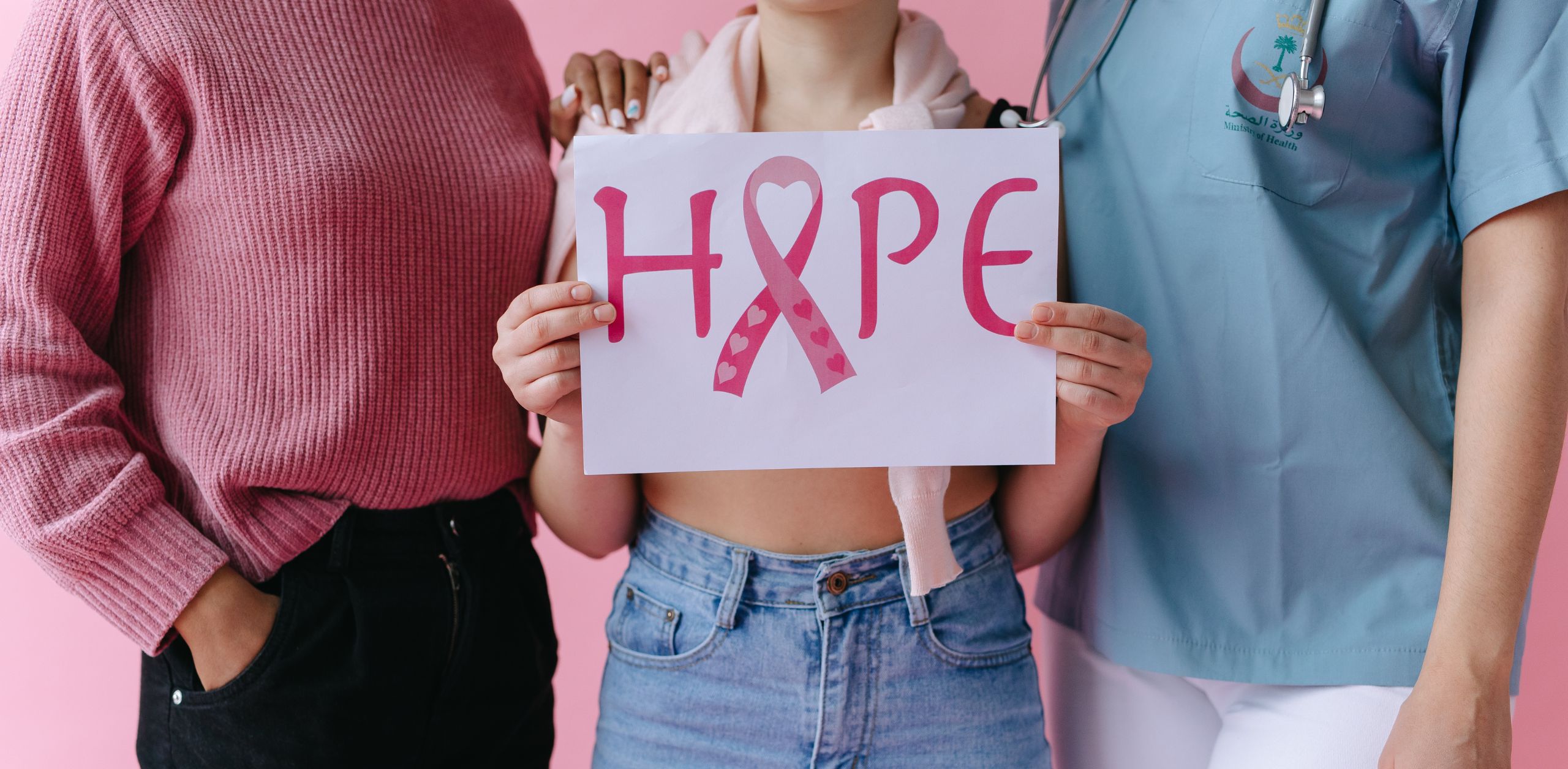 Young woman holding a “HOPE” sign with a pink breast cancer ribbon, standing between two supportive adults—one in pink and one in medical scrubs—symbolizing family and medical support during breast cancer recovery.