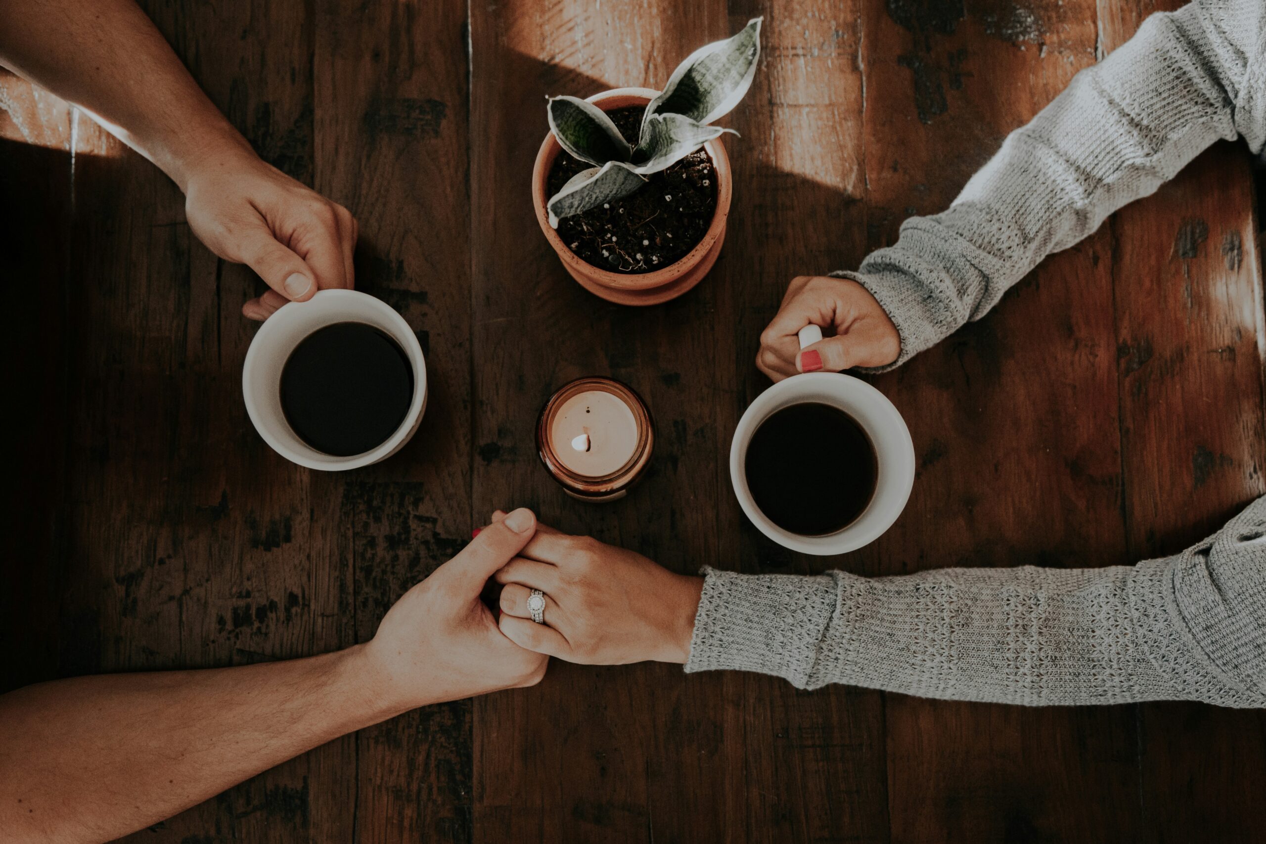 Two people holding hands over a wooden table with coffee mugs, a candle, and a plant, symbolizing love, support, and connection during a difficult time like cancer and feelings of guilt.
